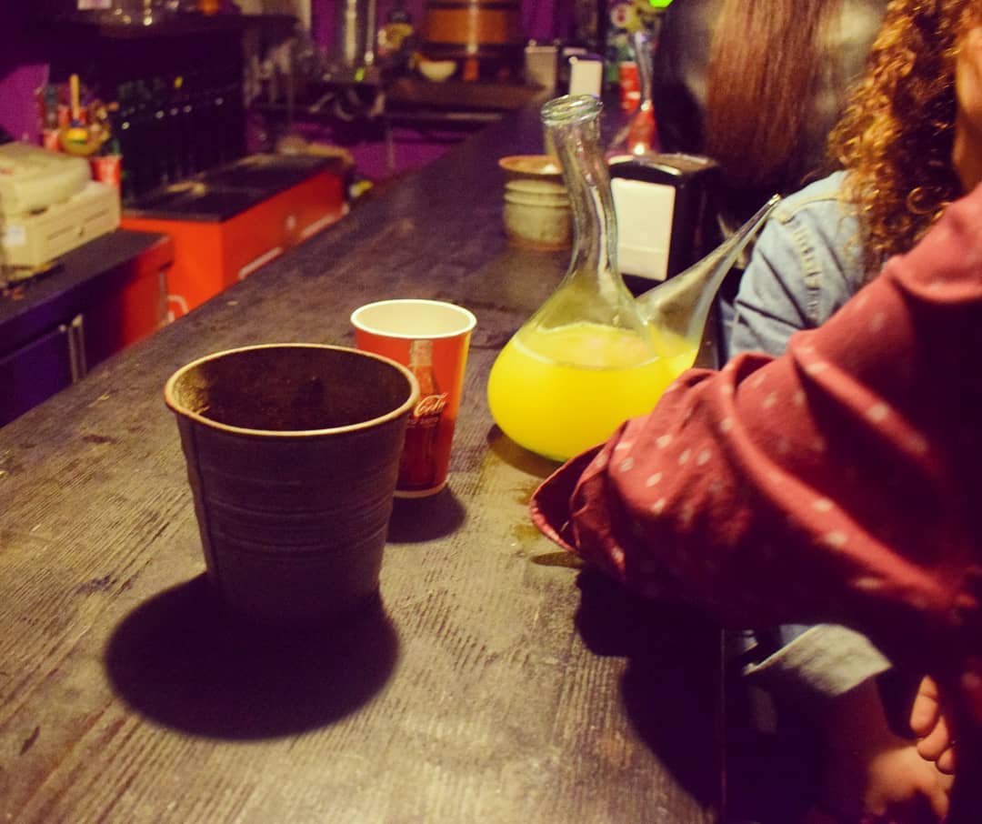 Fotografía da barra do Rincón do porrón. Enriba, un porrón, un vaso e un servilleteiro. Vense os codos de varios clientes apoiados na barra, conversando, na marxe dereita.