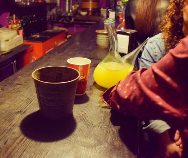 Fotografía da barra do Rincón do porrón. Enriba, un porrón, un vaso e un servilleteiro. Vense os codos de varios clientes apoiados na barra, conversando, na marxe dereita.